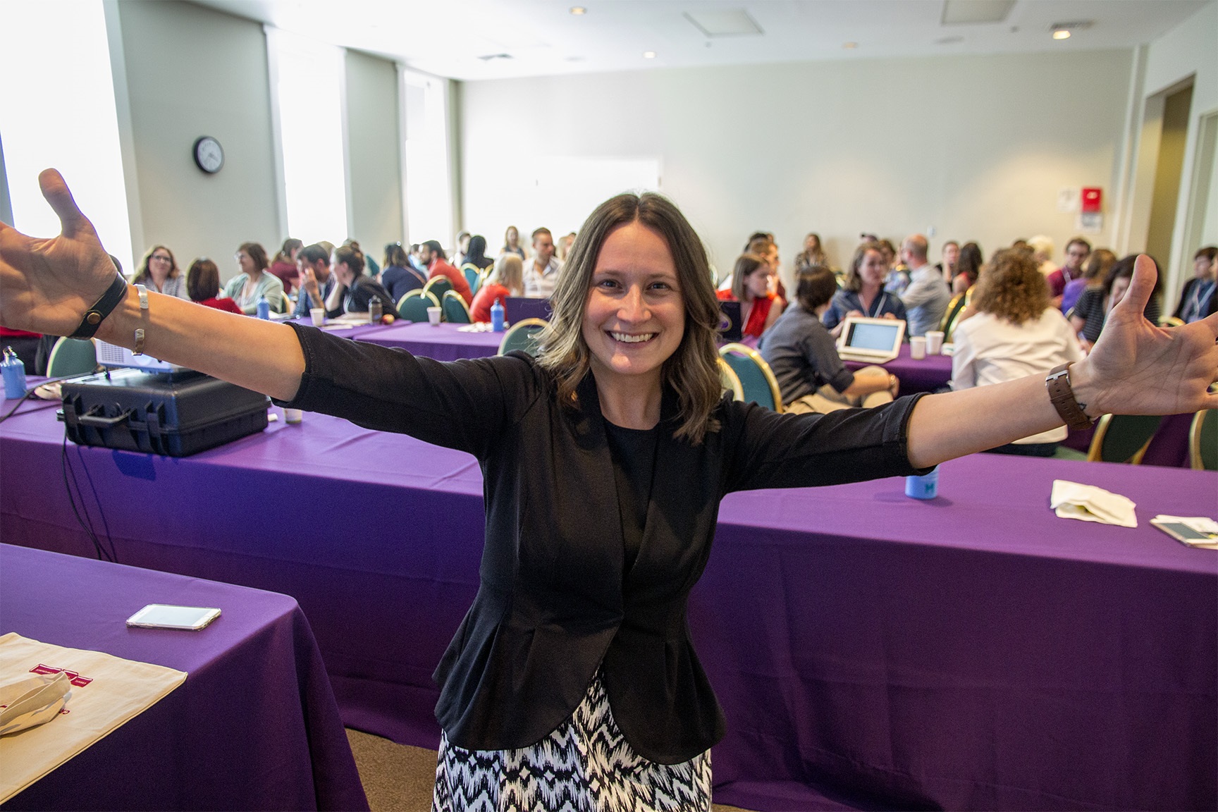 A woman leading a meeting smiles and extends her arms to the camera in front of the room full of meeting participants