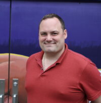 A man in a red shirt stands in front of a purple wall