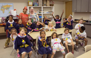 Group of children sit around a table in performance costumes making silly faces at the camera. Their two teachers stand behind