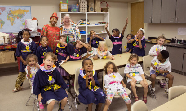 Group of children sit around a table in performance costumes making silly faces at the camera. Their two teachers stand behind
