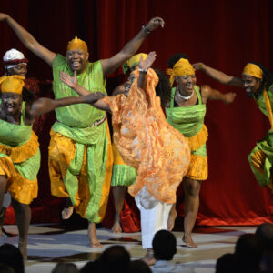 A group of dancers perform African dance in traditional costumes and bare feet on stage with a red curtain behind