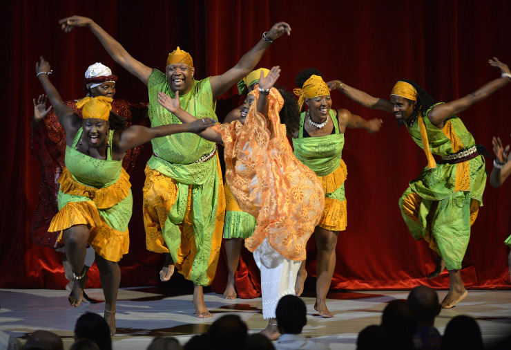 A group of dancers perform African dance in traditional costumes and bare feet on stage with a red curtain behind