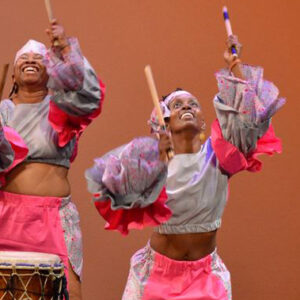 Two women in bright costumes hold drum sticks in the air during a performance