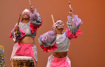 Two women in bright costumes hold drum sticks in the air during a performance