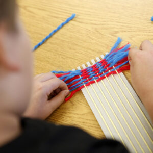 Looking over the shoulder of a child who is weaving pieces of wool onto a simple craft loom