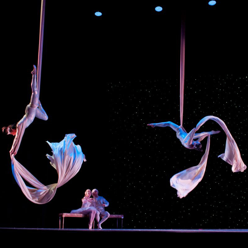 Four dancers perform on stage. Two aerial dancers hang from silks, two other dancers sit on a bench on stage and watch them