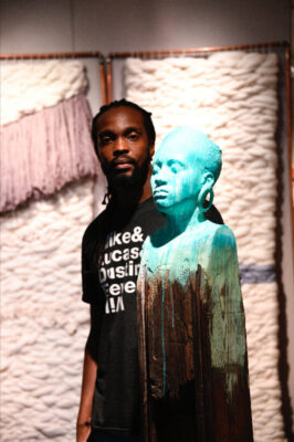 A male artist stands behind a sculpture of a female bust with bright paint dripping down it