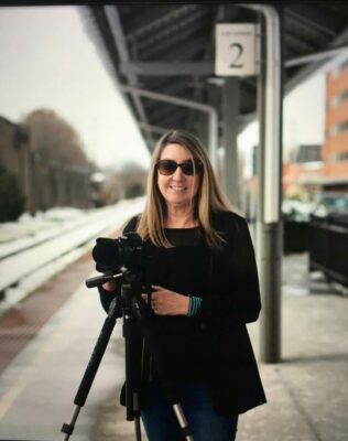 A woman stands on a train platform with a camera on a tripod smiling