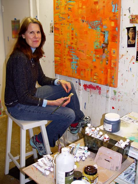 A woman sits on a stool in front of a painting holding a paintbrush and smiling at the camera