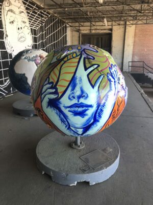 Globe painted with a monotone face cradled in two hands surrounded by bright tropical leaves