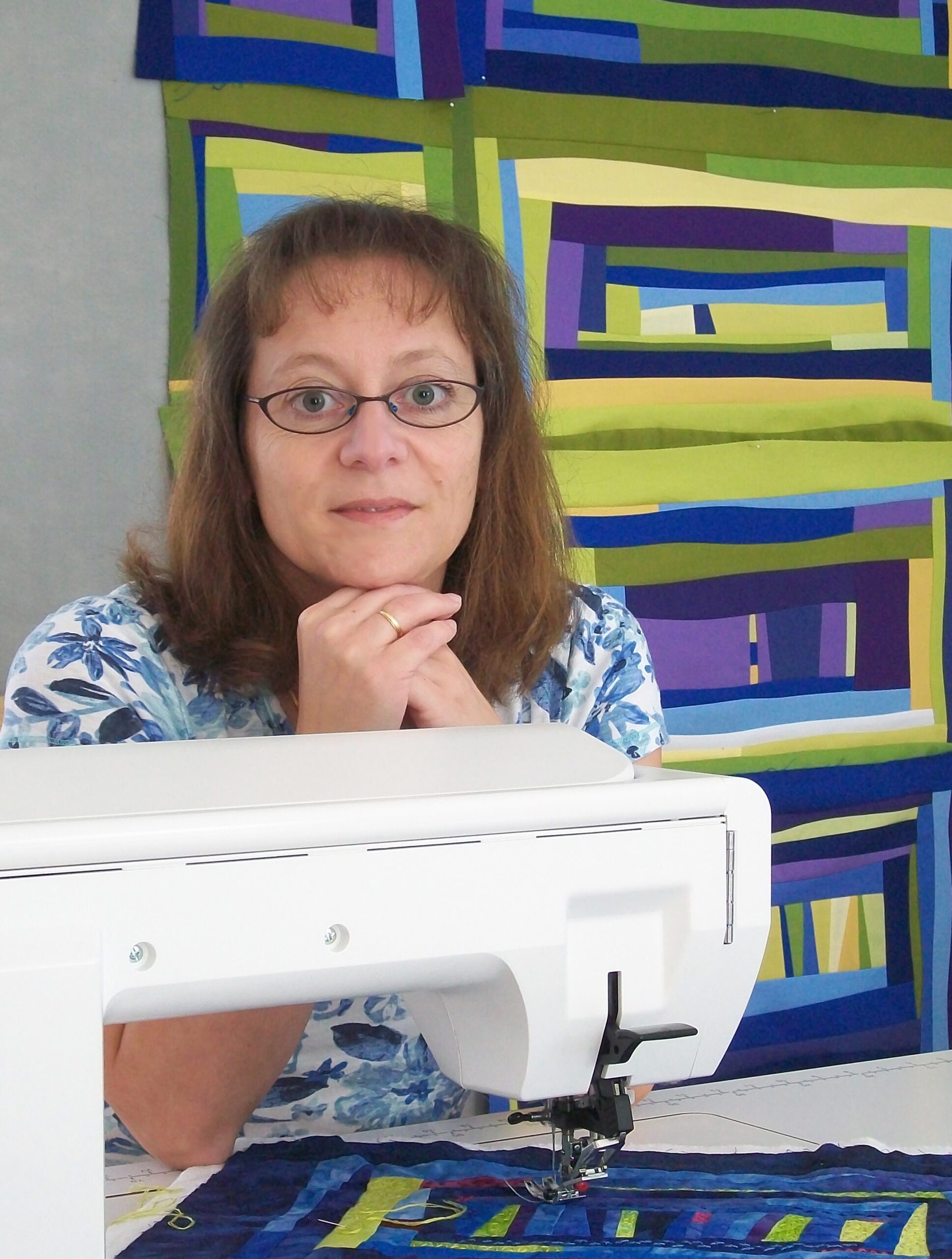 A woman sits behind a sewing machine with a project on it. There is a tapestry on the wall behind her that is very similar to the project she is working on