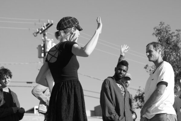 Black and white image taken from below eye level of a woman addressing a group of dancers outside during a rehearsal