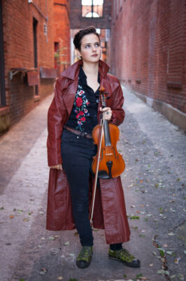 A woman stands in a brick alleyway holding a violin and bow. She is looking at the sky