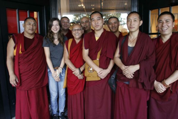 A woman in street clothes stands among a group of Buddhist monks in traditional robes. All are looking at the camera