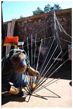 An artist kneels on the ground outside welding metal rods together into a geometric sculpture.