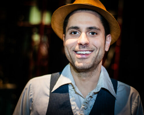 A man in a vest, shirt and straw hat smiles at the camera