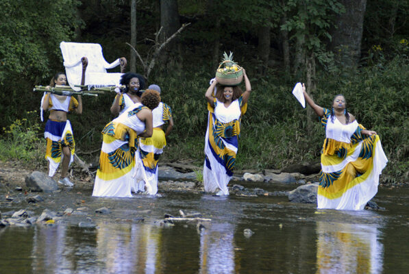 Six women in flowing, brightly patterned dresses wade in a river. They move in a procession. Several women hoist a white upholstered chair over their shoulders, and a smiling woman balances a basket on her head.