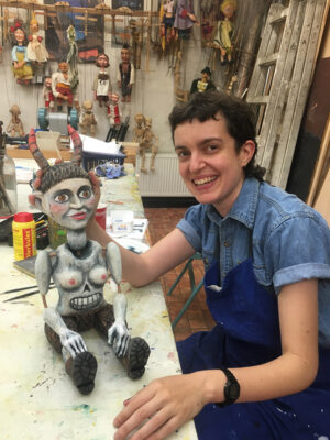 A woman in an apron is seated in a workshop. She props up a carved marionette puppet that sits on a work bench.